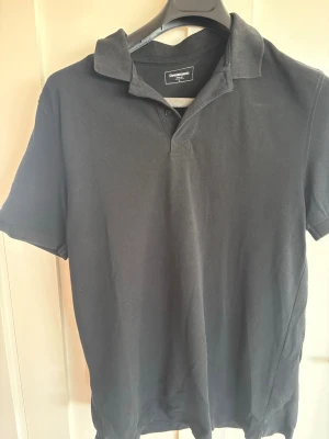 Svart pikétröja från Dressmann - Klassisk svart pikétröja från Dressmann med korta ärmar och krage. Tröjan har knappar vid halsen och är tillverkad i ett mjukt bomullsmaterial. Perfekt till jeans eller shorts för en enkel och stilren look.