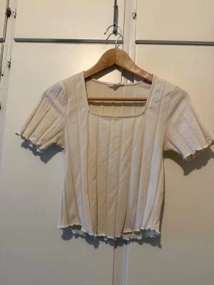 Beige ribbad topp med kort ärm - Snygg beige topp med ribbad struktur och korta ärmar. Toppen har fyrkantig ringning och vågig kant vid ärmslut och nederkant. Perfekt att styla med jeans eller kjol för en trendig look.