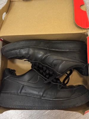 Svarta Nike Air Force 1 sneakers - Klassiska helsvarta Nike Air Force 1 sneakers i lågt utförande. Skorna har en robust sula, perforerad tåbox och ikoniska swoosh-loggan på sidan. Tillverkade i läder med svarta skosnören och diskret AIR-detalj på sulan.