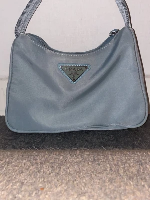 Ljusblå axelremsväska i satin - Snygg ljusblå handväska i satin med kort axelrem och dragkedja. Väskan har ett elegant mönstrat innerfoder i beige och silverdetaljer på dragkedjan. Perfekt för dig som gillar en stilren och trendig look.