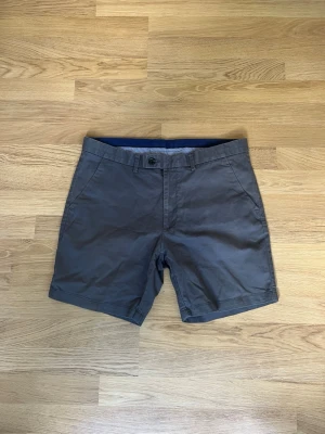 Gråa bomullsshorts med knapp - Snygga grå shorts i bomull med klassisk passform. De har knapp och dragkedja framtill, bälteshällor och två sidofickor samt två bakfickor med knapp. Perfekta för varma dagar och enkel att matcha med olika stilar.