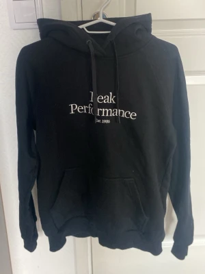 Svart hoodie Peak Performance S - Svart hoodie från Peak Performance med vit logga och text på bröstet. Klassisk modell med huva, dragsnören och stor magficka framtill. Tillverkad i mjukt material, perfekt för en avslappnad stil.