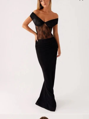 Svart långklänning med spetsdetaljer - Elegant svart långklänning med offshoulder och transparent spets upptill. Klänningen har figurnära passform, omlott över bysten och lång, slät kjol. Spetsen har blommigt mönster och ger en sofistikerad look.