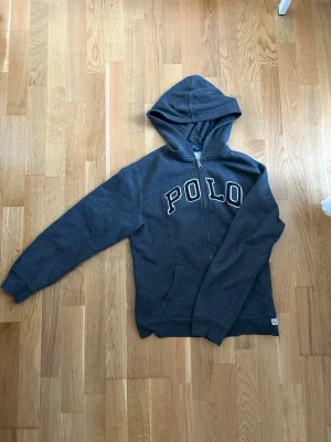 Mörkgrå hoodie med dragkedja från Polo - Snygg mörkgrå hoodie med dragkedja och  huva från Polo. Hoodien har vita bokstäver på bröstet och två fickor framtill. Perfekt för en avslappnad och cool stil.