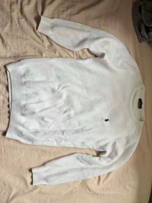 Vit stickad tröja från Polo Ralph Lauren - Klassisk vit stickad tröja från Polo Ralph Lauren med rund halsringning och diskret broderad logga på bröstet. Tröjan har lång ärm och ribbade muddar vid ärmslut och nederkant. Perfekt till lager på lager och enkel att matcha. Står M passar som S.