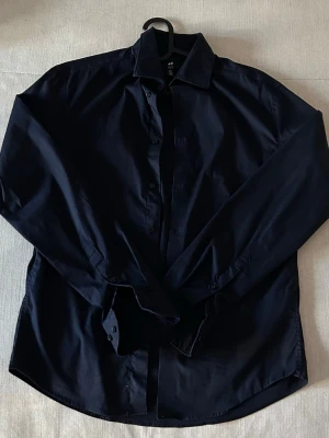 Mörkblå skjorta från H&M - Stilren mörkblå skjorta från H&M med klassisk krage och långa ärmar. Skjortan har knappar framtill och är tillverkad i ett mjukt bomullstyg. Perfekt för en clean och enkel look. Slim fit, skit snygg. Mörkblå.