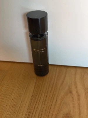 Zara Prima Parte Lychee & Oud parfym - En stilren parfymflaska från Zara med doften Prima Parte Lychee & Oud. Flaskan är mörkbrun och cylinderformad med svart lock. Texten på flaskan är i guld. Perfekt för dig som gillar lyxiga och unika doftkombinationer.