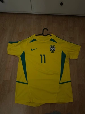Brasilien fotbollströja Ronaldinho #11 - Gul Brasilien fotbollströja från Nike med gröna detaljer och nummer 11. På bröstet finns Brasiliens landslagsemblem och på ryggen står det 'RONALDINHO' samt nummer 11. Perfekt för dig som älskar fotboll och vill stötta Brasilien.