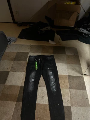 Svarta Dsquared2 jeans - Säljer ett par svarta jeans från Dsquared2 med slitningar, vita färgstänk och en grön nyckelring med logga. Jeansen har dragkedja framtill och smal passform. Perfekta för dig som gillar streetwear och unika detaljer.
