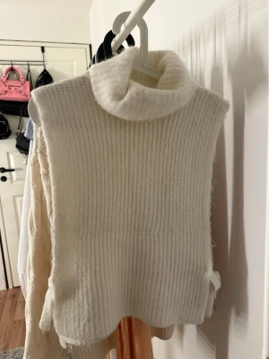 Vit stickad polokrage väst - Säljer en vit stickad väst med hög polokrage. Västen är ärmlös och har en mjuk, ribbad struktur. Materialet känns fluffigt och mysigt.