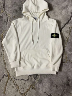 Vit hoodie från Stone Island - Säljer en stilren vit hoodie från Stone Island med klassisk huva och känguruficka. Tröjan har den ikoniska Stone Island-loggan på vänster ärm och ribbade muddar. Perfekt för dig som gillar streetwear och enkel design.