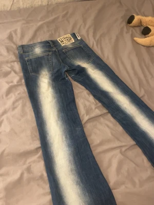 Blåa Cheap Monday jeans med blekta detaljer - Säljer ett par blåa jeans från Cheap Monday med tydliga blekta partier längs benen och bakfickor. Jeansen har raka ben och en vit patch med logga bak i midjan. Klassisk denimlook med en edgy twist. Säljer för de inte passar. Hålet syns inte så mycket och det är enkelt att sy 
