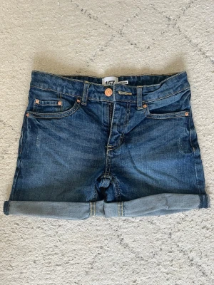 Blå denimshorts med uppvik - Snygga blå denimshorts med klassisk femficksmodell och uppvikta benslut. Shortsen har normal passform, bälteshällor och knappgylf. Perfekta för varma dagar och enkla att matcha med olika stilar.