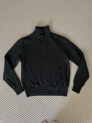 Ralph Lauren Svart stickad tröja med knappar - Mysig svart/mörkgrå stickad polotröja med ribbade muddar och bred ribbad nederkant. Tröjan har hög krage med två knappar framtill för extra stil och värme. Perfekt för kalla dagar och enkel att matcha med olika outfits.