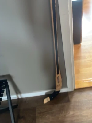 Träklubba för hockey - En klassisk hockeyklubba i trä med naturfärgad och svart design. Klubban har ett böjt blad och är perfekt för dig som gillar att spela hockey eller samla på sportprylar. Passar både till spel utomhus och inomhus.