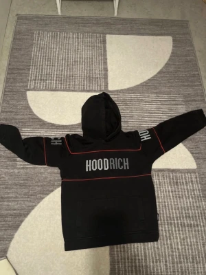 Svart HOODRICH hoodie med röda detaljer - Säljer en svart hoodie från HOODRICH med tydlig logga på bröstet och text på ärmarna. Hoodien har röda linjer som detaljer och en stor magficka framtill. Klassisk huva och avslappnad passform, perfekt för en streetwear-look.