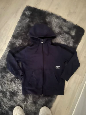 Mörkblå hoodie med dragkedja EA7 - Snygg mörkblå hoodie med dragkedja framtill och stor huva. Framtill finns två fickor och en liten EA7-logga på bröstet. Hoodien har långa ärmar och ribbade muddar vid ärmslut och nederkant. Perfekt för en avslappnad och cool stil.