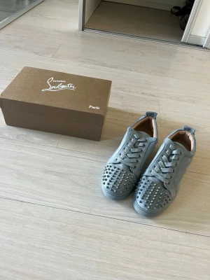 Gråa sneakers med nitar Louboutin - Säljer ett par gråa sneakers från Christian Louboutin i mocka med coola silverfärgade nitar på tån. Skorna har klassisk låg profil, rund tå och matchande grå snörning. Insidan är beige och sulan är ton-i-ton. Perfekta för dig som vill sticka ut.