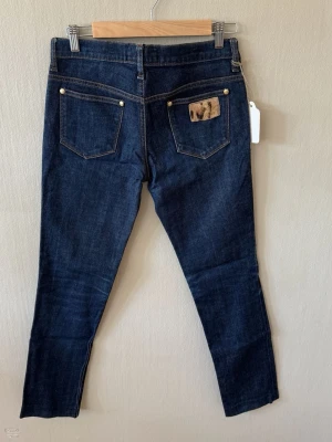 Love Moschino Mörkblå skinny jeans med patch - Snygga mörkblå skinny jeans från Love Moschino med klassisk femficksdesign och guldfärgade nitar. På ena bakfickan finns en unik patchdetalj i beige/brunt. Jeansen har normal midja och är tillverkade i stretchigt denimtyg för skön passform.0102