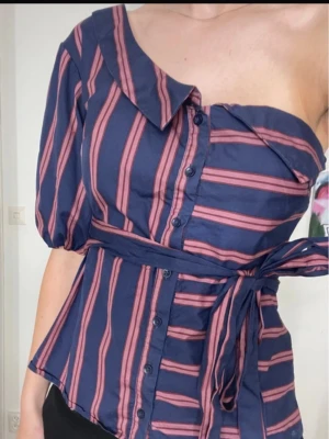 Asymmetrisk blus med bälte - Säljer en unik och trendig asymmetrisk blus med marinblå bas och rosa/röda ränder. Blusen har en axel bar och en puffärm, knappar framtill samt ett matchande knytbälte i midjan. Perfekt för både vardag och fest! 