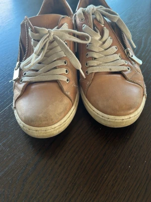 Bruna Charlotte of Sweden skor - Snygga bruna sneakers i läder-look med vita sulor och beige skosnören. Skorna har dragkedja på sidan för extra stil och enkel påtagning. Klassisk låg modell som passar till många outfits. Perfekta för dig som gillar en clean och stilren vibe.