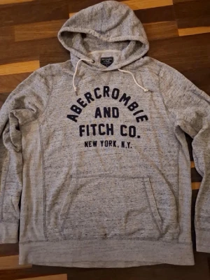 Grå hoodie från Abercrombie & Fitch - Snygg gråmelerad hoodie från Abercrombie & Fitch med svart tryck framtill och texten 'Abercrombie and Fitch Co. New York, N.Y.'. Klassisk känguruficka och justerbar huva med snören. Mjuk bomullsblandning som känns skön mot huden.