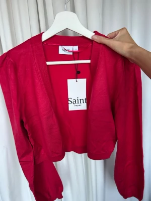 Röd cropped kofta från Saint Tropez - Snygg röd cropped kofta från Saint Tropez med långa puffärmar och ribbade muddar. Koftan har öppen front och är tillverkad i ett mjukt, tunt stickat material. Perfekt för att lägga till färg i din outfit.