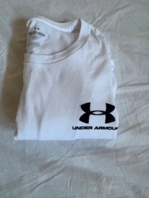 Vit Under Armour t-shirt - Vit t-shirt från Under Armour med svart logga och text på. Den har använts en gång då den var för stor för mig, det står att den är xs men den sitter som en s storlek.