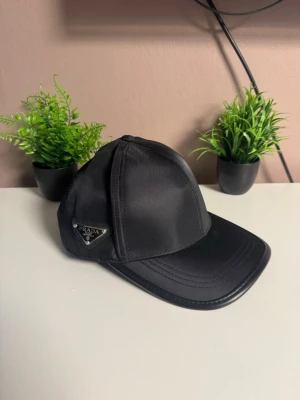 Svart keps från Prada i nylon - Snygg svart keps från Prada med klassisk böjd skärm och ikonisk metallogo på sidan. Kepsen är tillverkad i glansigt nylonmaterial som ger en exklusiv känsla och passar perfekt till streetwear eller en stilren look.