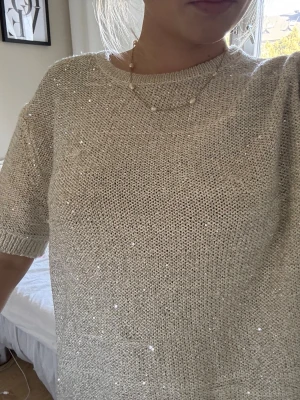 Beige glittrig stickad topp - Säljer en beige stickad topp med korta ärmar. Toppen har ett glittrigt material som ger en snygg touch och är perfekt för dig som vill ha något extra i garderoben. Passformen är loose och toppen är superenkel att styla till olika outfits.
