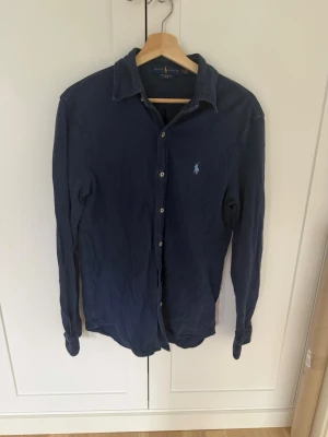 Mörkblå skjorta från Ralph Lauren - Klassisk mörkblå skjorta från Ralph Lauren i lätt mesh-bomull. Skjortan har långa ärmar, knappar framtill och den ikoniska broderade loggan på bröstet. Perfekt till jeans eller chinos för en clean och stilren look.