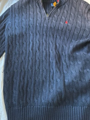 Mörkblå kabelstickad tröja Polo Ralph Lauren - Kabelstickad mörkblå tröja från Polo Ralph Lauren med halv dragkedja och röd broderad logga på bröstet. Tröjan har ribbade muddar och krage med röd rand på insidan. Perfekt för dig som gillar klassisk och stilren design.