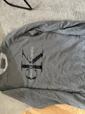 Grå sweatshirt från Calvin Klein - Snygg grå sweatshirt från Calvin Klein Jeans med stort CK-logotyptryck på bröstet. Tröjan har rund halsringning och långa ärmar. Perfekt för en avslappnad och trendig look. (Inte säker på storlek men passar ”s”)