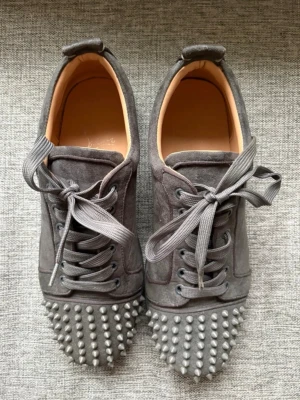 Gråa Louboutin skor med nitar - Grå sneakers från Christian Louboutin i mocka med silverfärgade nitar på tån. Skorna har röda sulor och matchande grå skosnören. Saknar en spik på vardera tå, därav priset. Storlek 44! Tillbehör medföljer som skohorn, kort, påsar.