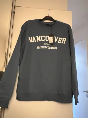 Blå sweatshirt Vancouver REACTION XL - Blå sweatshirt från REACTION i storlek XL med trycket 'VANCOUVER BRITISH COLUMBIA' i vitt framtill. Tröjan har rund halsringning, ribbade muddar vid ärmslut och nederkant samt en avslappnad passform. Perfekt för en chill och casual stil.
