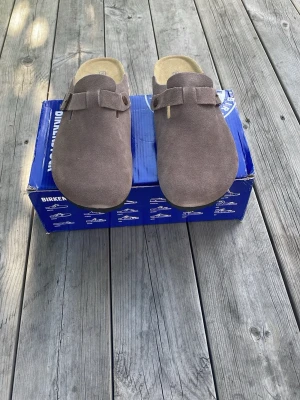 Bruna Birkenstock tofflor i mocka - Säljer ett par Birkenstock tofflor i mjuk mocka med justerbart spänne och korksula. Klassisk slip-in modell med präglad logga på sidan och bekväm innersula. Perfekta för chill dagar hemma eller ute. Färgen är dk grey men jag skulle säga att den är mer brunaktig. Tofflorna är i perfekt skick och är bara testade, aldrig använda utomhus. Bara boxen som är sliten. Skriv vid funderingar 😊