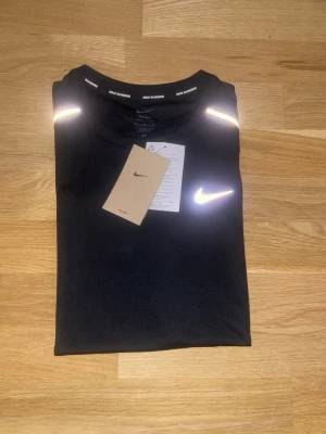 Svart löpartröja från Nike - Svart löpartröja från Nike med reflekterande detaljer på axlarna och ett vitt Nike Swoosh på bröstet. Tillverkad i ett lätt och snabbtorkande material, perfekt för löpning och träning. Rund halsringning och diskret Nike Running-tryck på insidan.