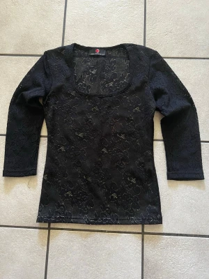 Svart spetsblus storlek S - Snygg svart blus i spets med blommigt mönster och trekvartsärm. Toppen har en rundad, djup urringning och är genombruten i materialet vilket ger en elegant look. Perfekt att styla med jeans eller kjol.