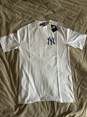 Vit New York Yankees t-shirt popcorn - Säljer en vit t-shirt med New York Yankees-logga på bröstet och en stor popcorn-illustration med Yankees-keps på ryggen. T-shirten har korta ärmar och är gjord i mjuk bomull. Perfekt för dig som gillar streetwear och sportiga vibes. !!! XS OVERSIZED FIT!!!