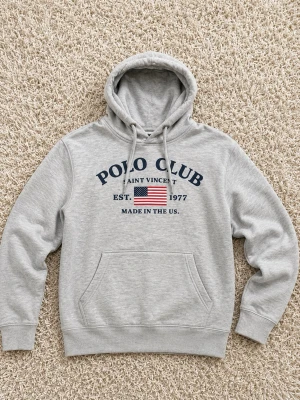 Polo club Hildur - Snygg grå hoodie med huva och dragsnören. På bröstet står det 'POLO CLUB SAINT VINCENT' och 'EST. 1977' tillsammans med en amerikansk flagga. Stor magficka framtill och ribbade muddar vid ärmar och nederkant. Perfekt för en avslappnad streetwear-look.