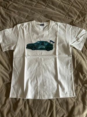Vit Stussy t-shirt med biltryck - Säljer en vit t-shirt från Stussy med ett stort tryck av en mörkgrön bil på bröstet. T-shirten har klassisk passform, rund hals och är kortärmad. Perfekt för dig som gillar streetwear och coola prints. Storlek L men passar mer som M