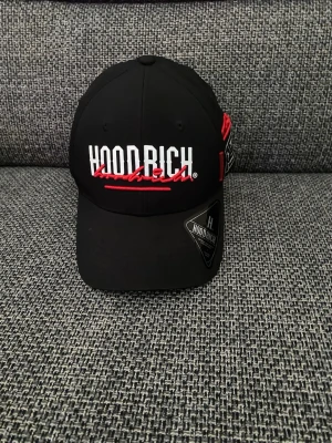 Hoodrich Cap - Pratar svenska också. This is a black Hoodrich branded cap featuring white and red embroidery on the front. The inside of the cap has branded taping and a label with product details. The cap appears to be new, with a sticker still attached to the brim never used because it was not my style. But no tags