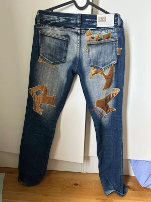 Blå bootcut lowrise jeans med gulda detaljer från AnyWay - Väldigt lowrise. Unika blå jeans med slitningar och stora guld patch-detaljer i graffiti-stil på baksidan. Jeansen har raka ben, låg midja och klassiska fyra fickor. Materialet är denim med en tvättad look och kontrastsömmar. Perfekta för dig som vill sticka ut med din stil. New york style