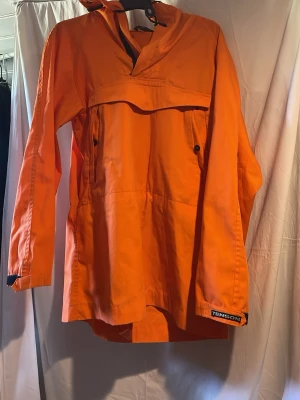 Orange regnjacka från Tenson - Snygg orange regnjacka från Tenson med huva och dragkedja vid halsen. Jackan har praktiska fickor med knappar och justerbara ärmslut. Perfekt för regniga dagar och outdoor-äventyr.