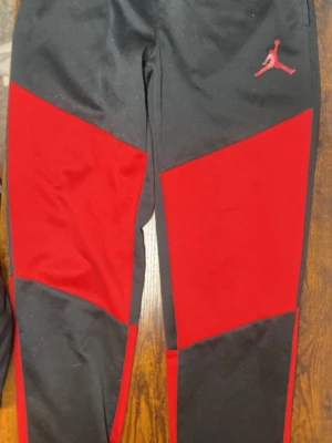 Jordan svart/röd byxa - Snygga träningsbyxor från Jordan i svart och rött med det klassiska Jumpman-loggan på vänster sida. Byxorna har dragsko i midjan för justerbar passform och är tillverkade i ett mjukt och bekvämt material. Perfekta för både träning och vardag.
