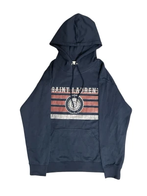 Saint Laurent hoodie - Snygg mörkblå hoodie med stor huva och dragsko. Framsidan har ett vintageinspirerat tryck med texten 'SAINT LAURENT' och grafiska ränder i vitt och rött. Praktisk magficka framtill. Perfekt för en avslappnad streetwear-look.