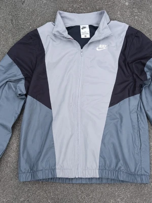 Grå och svart vindjacka från Nike - Snygg vindjacka från Nike i grått och svart med dragkedja framtill och hög krage. Jackan har en sportig look med färgblock och broderad Nike-logga på bröstet. Tillverkad i lätt och tåligt material, perfekt för blåsiga dagar.