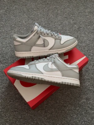 Nike Dunk Low grå/vit sneakers - Nike Dunk Low sneakers i stilren grå och vit färg. Skorna har klassisk låg siluett, perforerad tåbox och ikoniska swoosh-loggan på sidan. Ovandelen är i läder med matchande grå snören och vit mellansula. Perfekta för dig som gillar clean och tidlös streetstil.