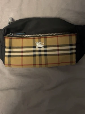Burbarry magväska - Snygg magväska i beige och svart med klassiskt rutigt mönster på framsidan och silverfärgad dragkedja. Väskan har justerbart band och en liten metall-logga framtill. Perfekt för att bära det viktigaste på ett stilrent sätt.