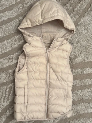 Beige dunväst med huva från ONLY - Snygg beige dunväst från ONLY med quiltad design och dragkedja framtill. Västen har en stor, mysig huva och två fickor på sidorna. Perfekt för lager på lager och ger en clean look till din outfit. Figursydd.